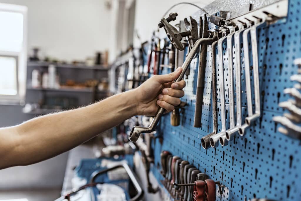 Photo of male mechanic reaching for Tools for Car arranged on the wall. Unrecognizable Person, a Caucasian male worker taking a tool from wall to repair a bicycle or car. Bike workshop interior. Young mechanic taking some tools in his shop. Motorcycle mechanic in his workshop taking a tool from his tool pane.