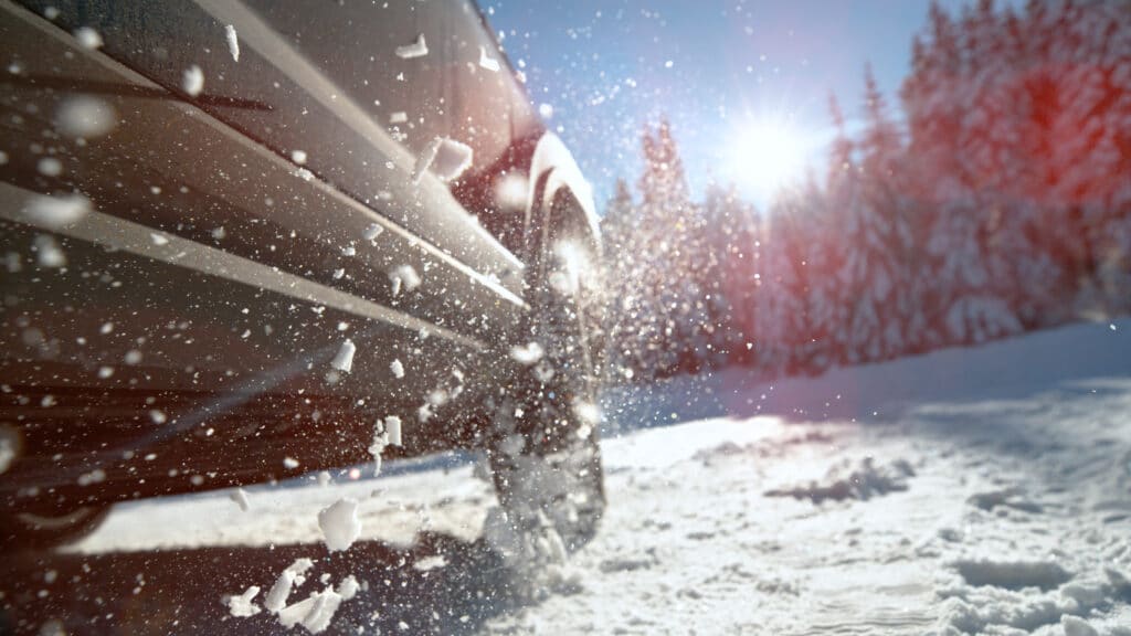 CLOSE UP, LENS FLARE, LOW ANGLE, DOF: Metallic blue car throws up chunks of snow while trying to drive along a snowy road in the idyllic Slovenian mountains. SUV struggles to get traction on icy road.