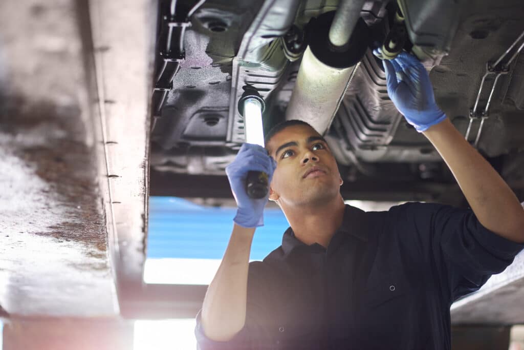 A young mechanic is working under a car in a garage repair shop. He is wearing blue overalls.  He is looking up with an inspection lamp to check the possible damage that has been sustained to the exhaust.